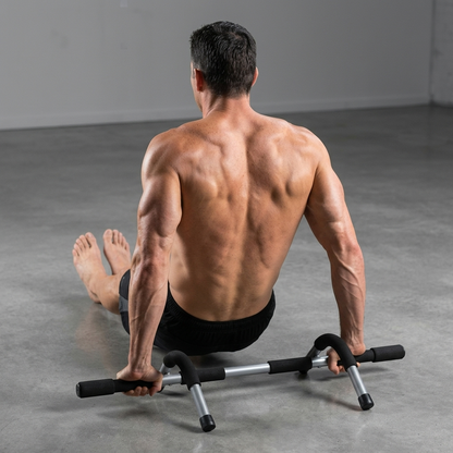 A shirtless man with a muscular back sits on a floor exercise door frame, viewed from behind. The black and silver door frame is positioned on a gray concrete floor in a minimalist studio setting.